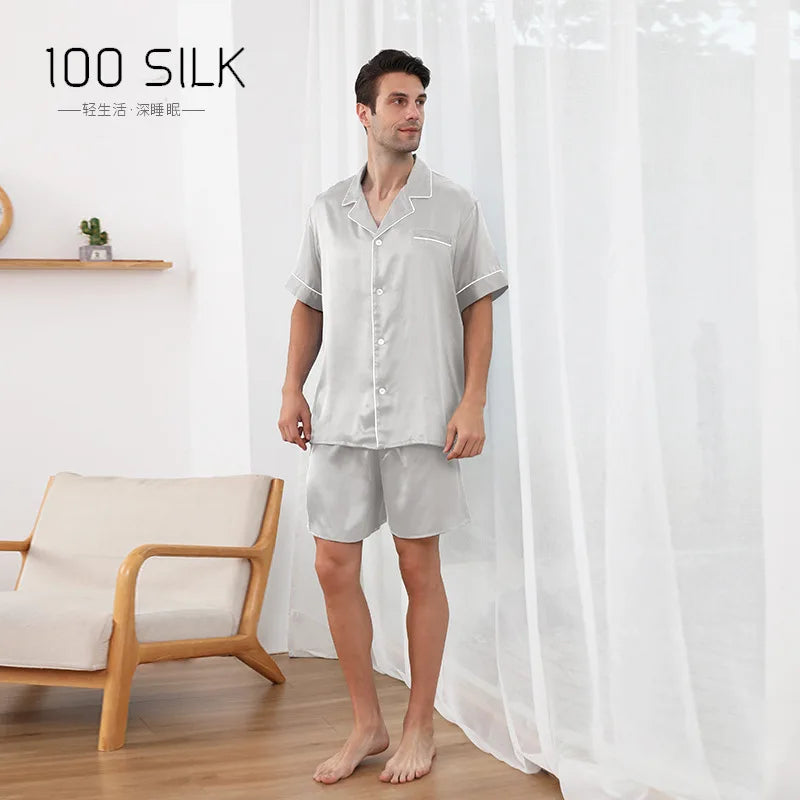 Man wearing a light gray silk pajama set standing in a room with a white curtain and wooden chair.
