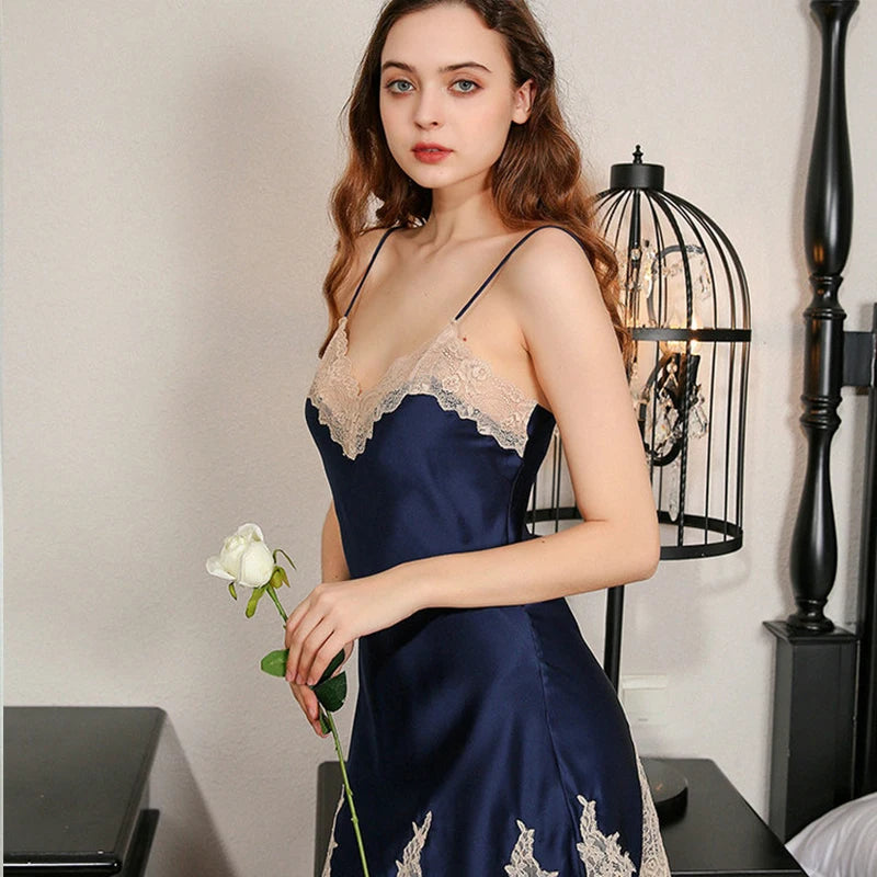 Woman in a navy blue satin nightgown with lace details holding a white rose in a bedroom setting.